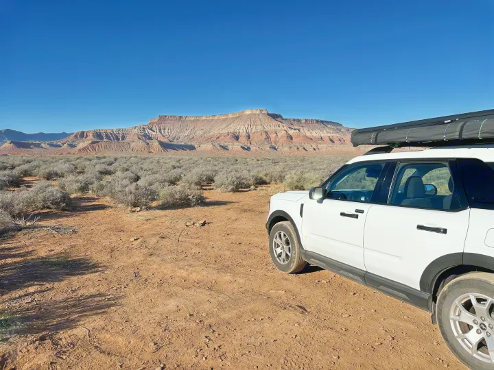 White SUV with a rooftop awning on a desert dirt road, backed by red rock mountains. Great for an RV rental outdoor adventure.. Ford Bronco Sport 2024