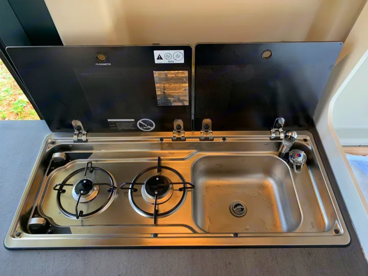 Compact RV rental kitchen features a Dometic two-burner gas hob and sink unit with open glass lids, ideal for camper cooking.. Sunlight Cliff 600 2023