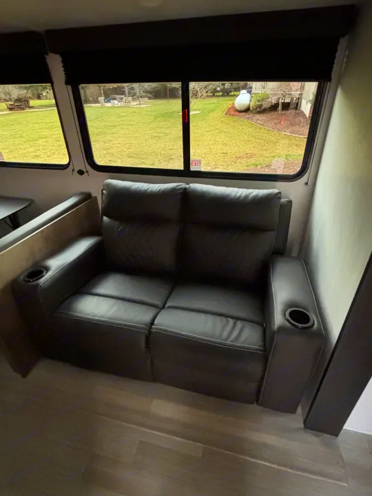 Inside this RV rental, find a dark leather recliner with cup holders next to large windows. An emergency exit sticker is visible.. Keystone Springdale 261BHC 2026