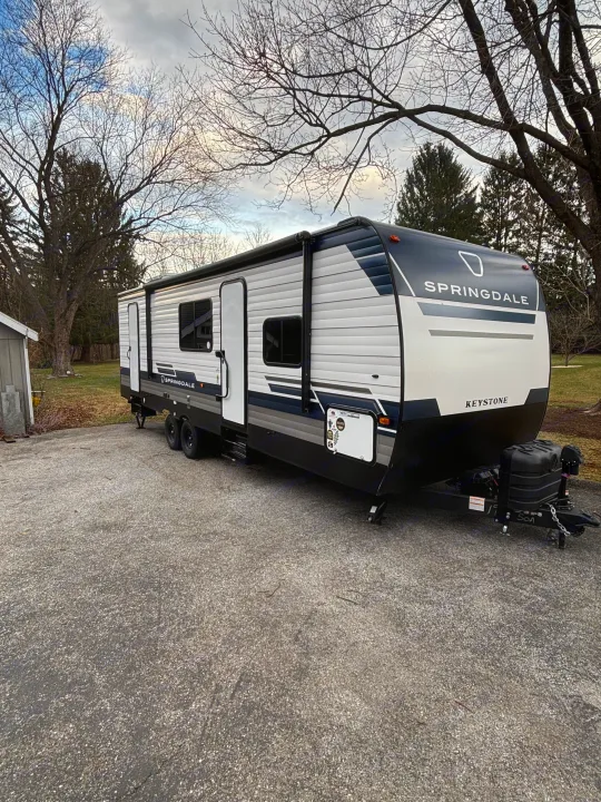 A white, black, and gray Springdale Keystone RV is parked on an asphalt driveway, ideal for an rv rental. Bare trees in the background.. Keystone Springdale 261BHC 2026