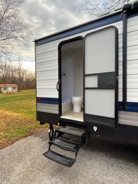 Explore this RV rental's open door, revealing a compact bathroom with a toilet and shower. Black steps lead to the entry, set in a natural area.. Keystone Springdale 261BHC 2026