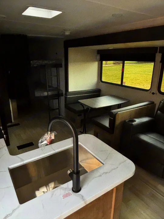 RV rental interior shows a modern kitchen sink, dinette, bunk beds, and a comfy recliner, with green grass visible outside.. Keystone Springdale 261BHC 2026