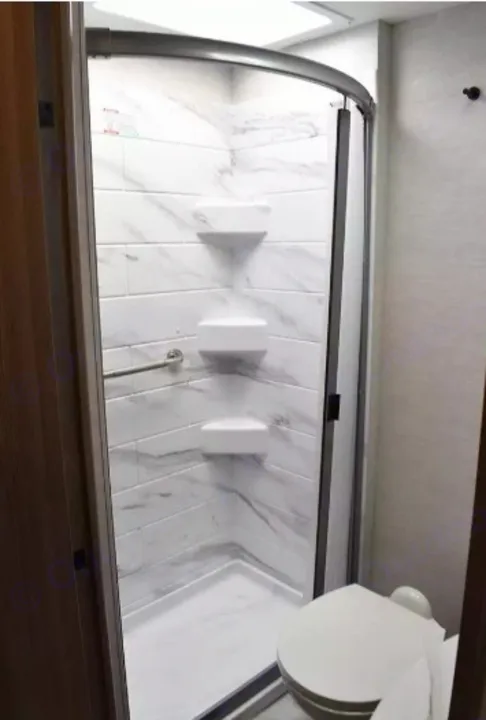 Compact RV rental bathroom with a curved shower featuring marbled walls, corner shelves, a grab bar, and a toilet.. Thor Eddie Bauer 2025