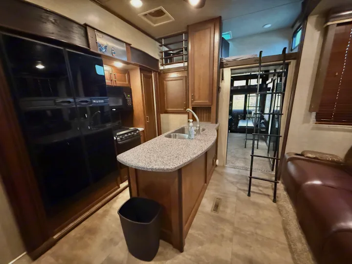 Spacious rv rental kitchen interior with an island, fridge, stove, and a ladder leading to an overhead bunk bed. A brown couch is also visible.. Keystone Fuzion 2016