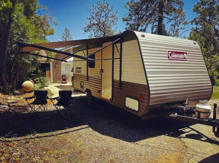 Ready for adventure: Coleman travel trailer RV rental with extended awning & chairs on a sunny gravel spot, surrounded by trees.. Dutchmen Coleman Lantern 2021
