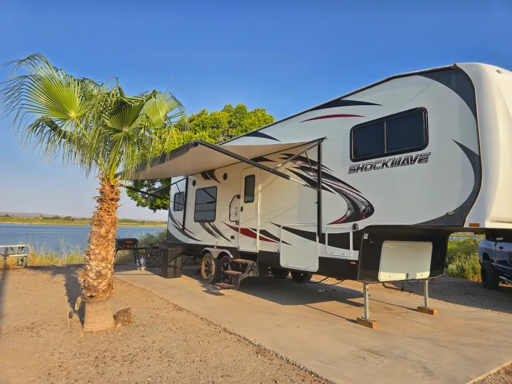 Experience this Shockwave fifth-wheel rv rental: A white RV with awning extended, parked by a scenic lake and palm tree.. Forest River Shockwave 2018