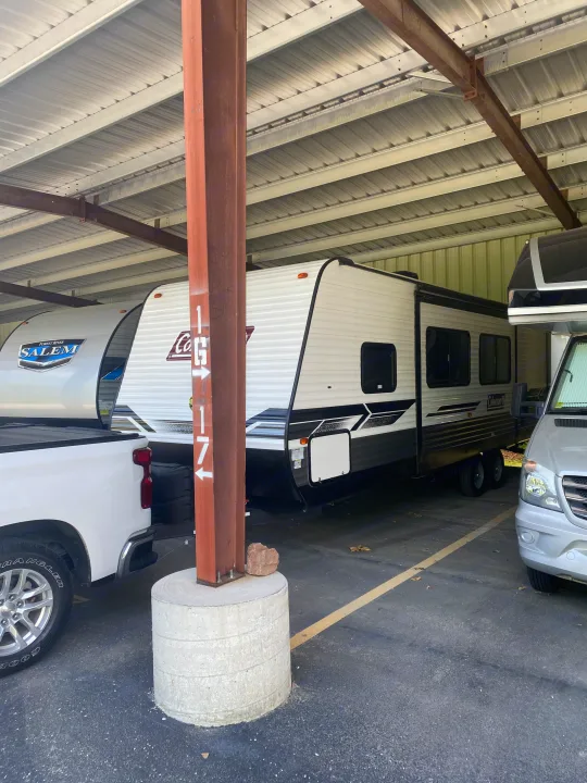 Diverse RVs (travel trailers, pickup, van) parked in a covered rv rental facility. A brown pillar with '1G17' is central, securing the structure.. Dutchmen Coleman Lantern LT 2023