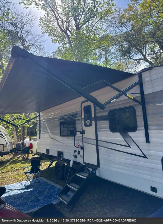 Access RV rental at a sunny campsite, with its awning extended over an outdoor rug and chair, ready for relaxation.. Winnebago Access 2024