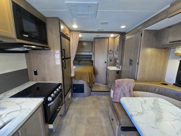 Modern RV rental interior: kitchen, dining booth, and bed area in light wood tones. Compact and well-equipped for travel.. Forest River Coachmen Prism 2020
