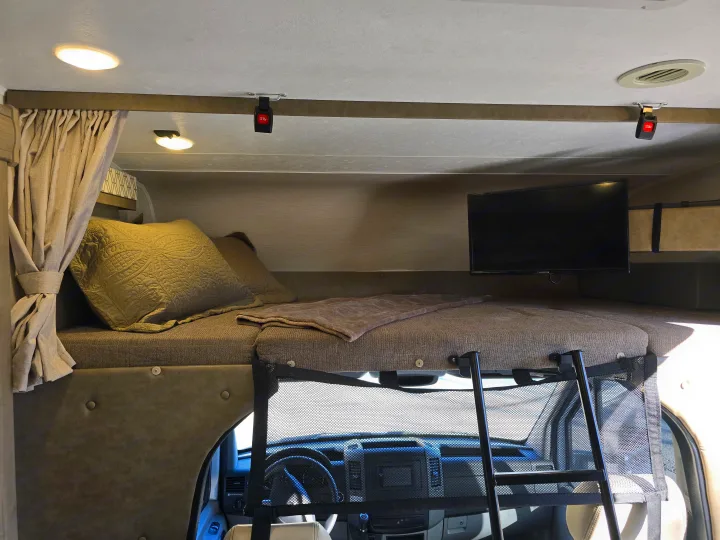 Overhead bunk bed in an rv rental, featuring a TV, safety net, pillows, blanket, ceiling lights, and view into the cab.. Forest River Coachmen Prism 2020