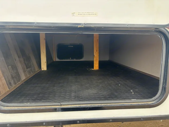 Spacious, empty RV rental exterior storage compartment with a rubber mat, wooden supports, and an interior access door.. KZ Escape 2018