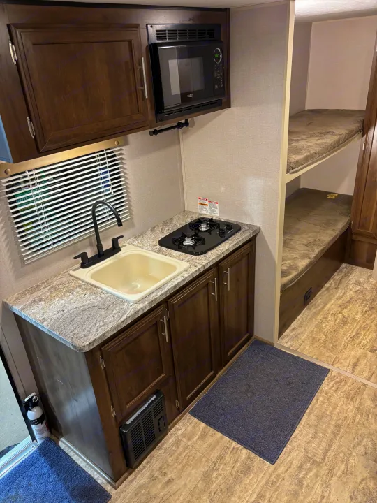 RV rental interior: a compact kitchen with dark cabinets, sink, two-burner stove, microwave, and twin bunk beds.. KZ Escape 2018