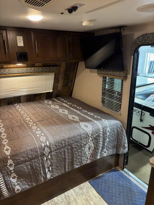 Cozy RV rental bedroom with patterned bed, wall-mounted TV, cabinets, and ceiling controls for comfortable travel.. KZ Escape 2018