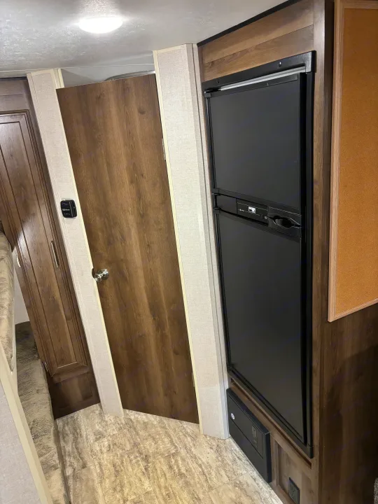 RV rental interior. Features a black refrigerator, dark wood door, light textured walls, and a wall-mounted thermostat.. KZ Escape 2018