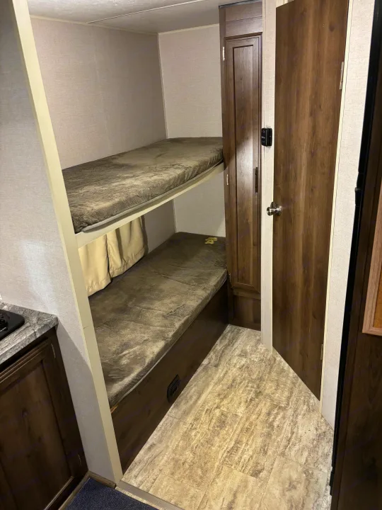 Inside an RV rental: two cozy bunk beds with fuzzy blankets, adjacent wooden storage, and a door with a silver knob. Textured walls & tiled floor.. KZ Escape 2018