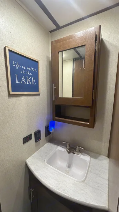 This **RV rental** bathroom vanity features a sink, mirror, a "life is better at the LAKE" sign, and RV controls. A blue night light adds ambiance.. Primetime Avenger 2019