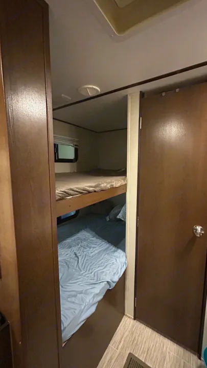 RV rental bunk bed sleeping area. Upper bed with grey bedding, lower with blue. Small window and closed door complete the RV interior.. Primetime Avenger 2019
