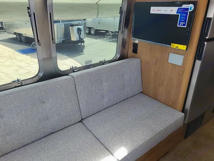 This RV rental interior features a cozy grey sofa, large window, and wall-mounted TV for comfortable travel.. Airstream Trade Wind 2025