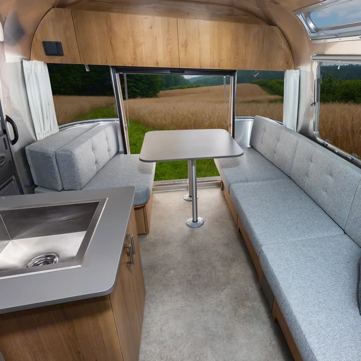 Cozy RV rental interior featuring light grey upholstered benches, a dining table, and a kitchen sink. Large windows offer scenic field views.. Airstream Trade Wind 2025
