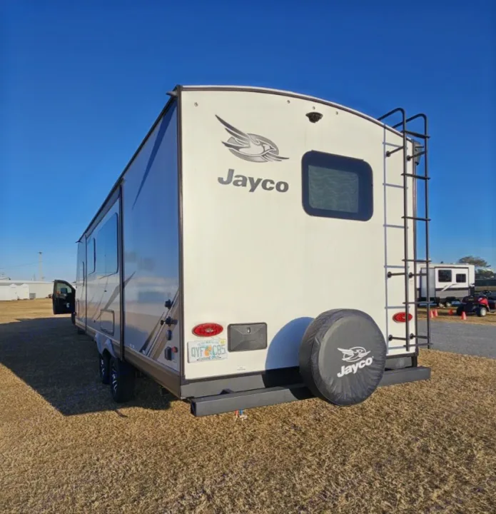 A white Jayco travel trailer RV rental, seen from the rear with a ladder and spare tire, parked outdoors under a blue sky.. Jayco White Hawk 2023