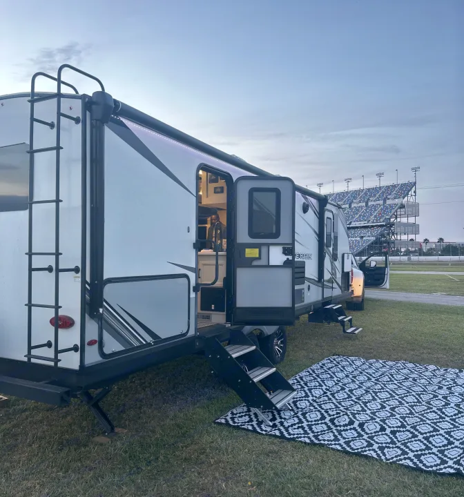 Dusk camping: RV rental beside a racetrack. Open door, person inside, mat & steps complete the cozy setup. Stadium lights visible.. Jayco White Hawk 2023