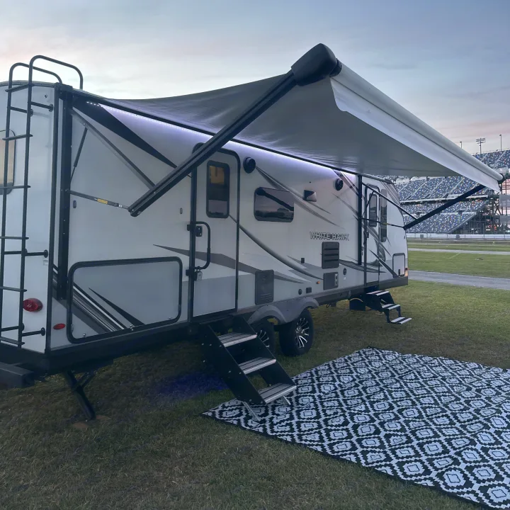 An RV rental with an extended, lit awning and outdoor mat set up on grass next to race track grandstands.. Jayco White Hawk 2023
