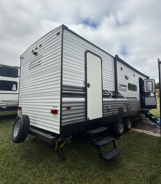 Explore the outdoors with this Coleman travel trailer rv rental, shown parked on grass with its steps down and a spare tire.. Coleman Lantern LT 26BH 2022