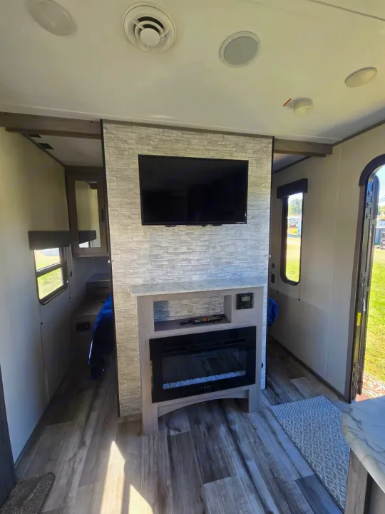 Cozy RV rental interior featuring a wall-mounted TV, electric fireplace, modern wood-grain floor, and bright natural light.. Coleman Lantern LT 26BH 2022