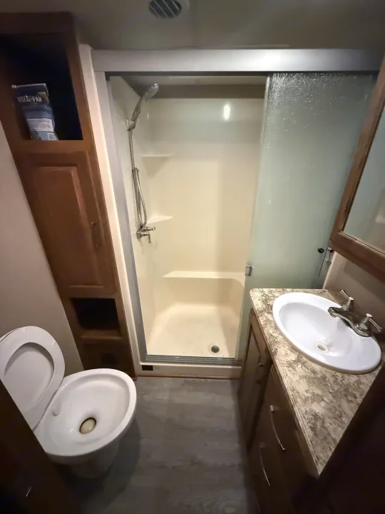 An RV rental bathroom showing a white toilet, compact shower stall with sliding door, and sink vanity with a speckled countertop.. Grand Design Reflection 2015