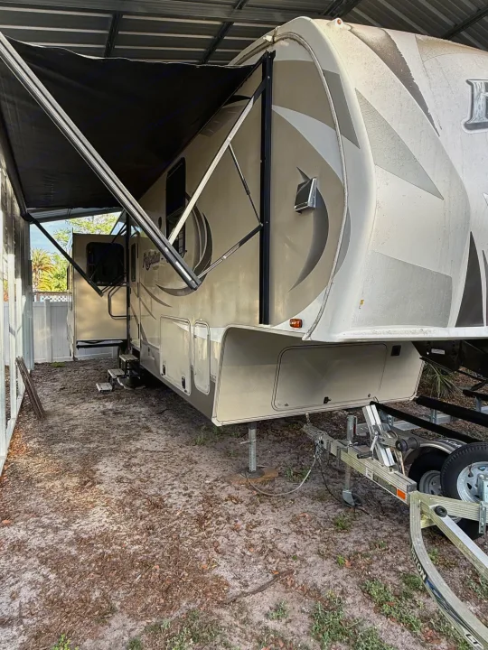 A beige RV rental with an extended black awning is parked under a metal shelter. Sandy ground, steps, and a boat trailer are visible.. Grand Design Reflection 2015