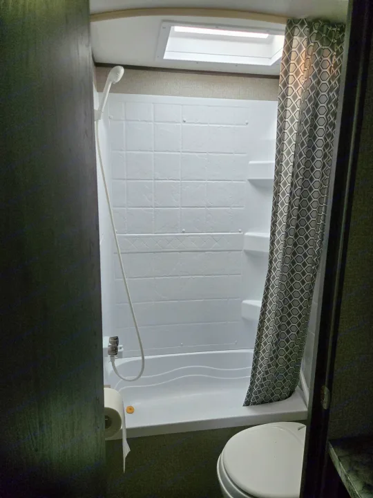This RV rental features a compact bathroom with a white shower/tub combo, shower head, toilet, and a geometric patterned shower curtain.. Jayco Jay Flight SLX 2021