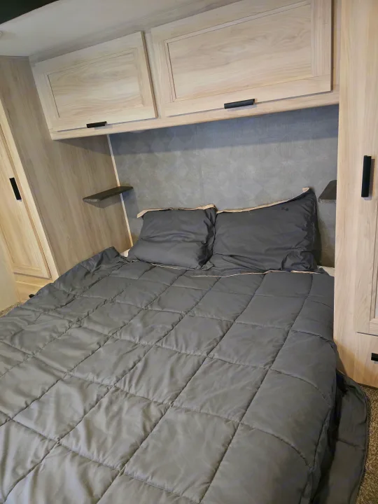 Inside a modern rv rental: a cozy bedroom with a quilted gray comforter, pillows, light wood cabinets, and shelving.. Forester LE 2023