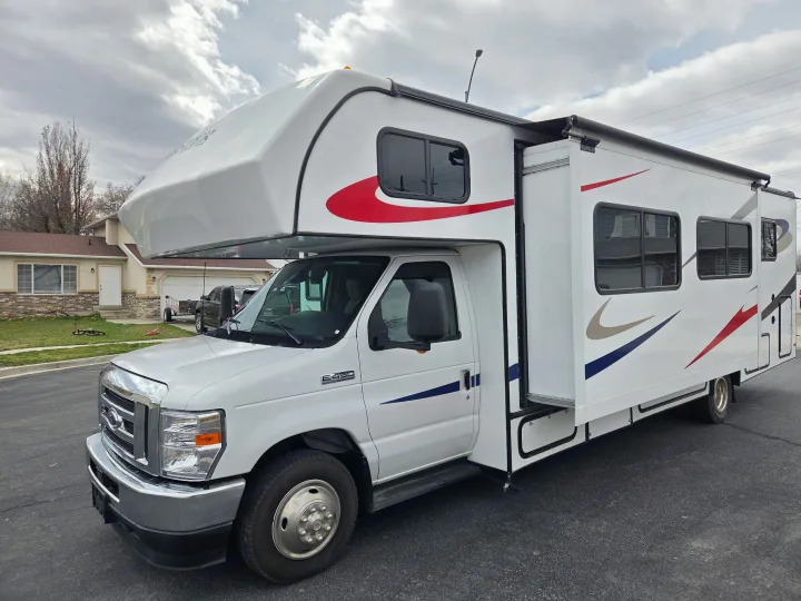 This white Ford E-450 Class C rv rental features red, blue, and tan decals, with a side slide-out extended.. Forester LE 2023