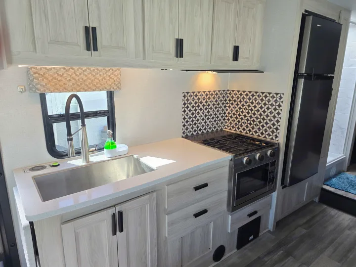 Modern rv rental kitchen with a stainless steel sink, stove, microwave, and refrigerator. Features light cabinets and a geometric backsplash.. Forester LE 2023