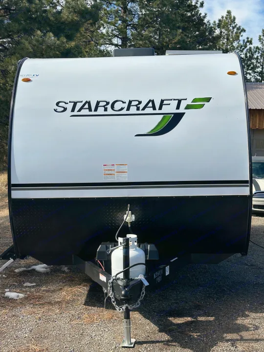 White and black Starcraft travel trailer for rv rental, featuring propane tanks and hitch. Pine trees in background.. Starcraft Autumn Ridge 2025