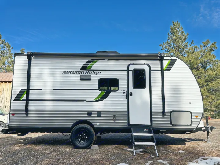 An Autumn Ridge travel trailer, perfect for rv rental, stands with white & gray siding, green accents, under a clear blue sky.. Starcraft Autumn Ridge 2025