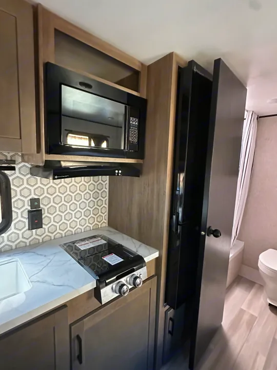 Compact rv rental kitchen with microwave, stovetop, sink, and backsplash, next to a glimpse of the bathroom.. Starcraft Autumn Ridge 2025