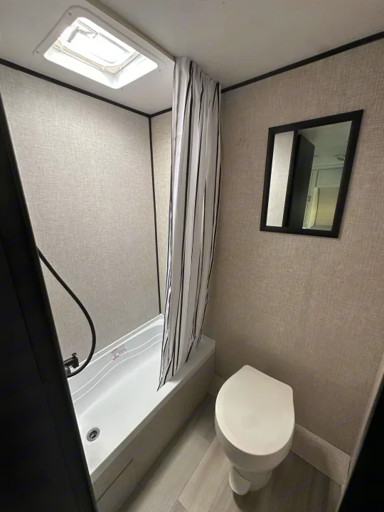Compact rv rental bathroom featuring a shower with a striped curtain, toilet, and mirror, all illuminated by a ceiling skylight.. Starcraft Autumn Ridge 2025