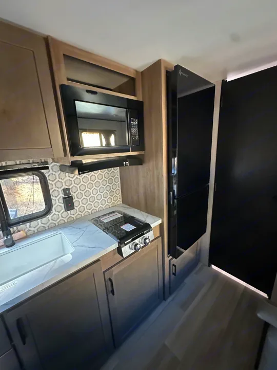 RV rental's modern kitchen features light cabinets, a black microwave, stovetop, sink, and tall refrigerator for comfort on the road.. Starcraft Autumn Ridge 2025