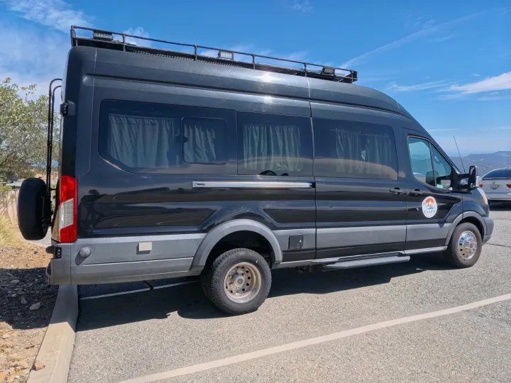 Black custom camper van from CLIFF, an RV rental, with a roof rack and rear spare tire, parked outdoors.. Ford Transit Custom 2015