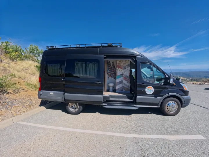 Black RV rental camper van with open side door revealing interior toilet & colorful wall, parked with mountain views.. Ford Transit Custom 2015