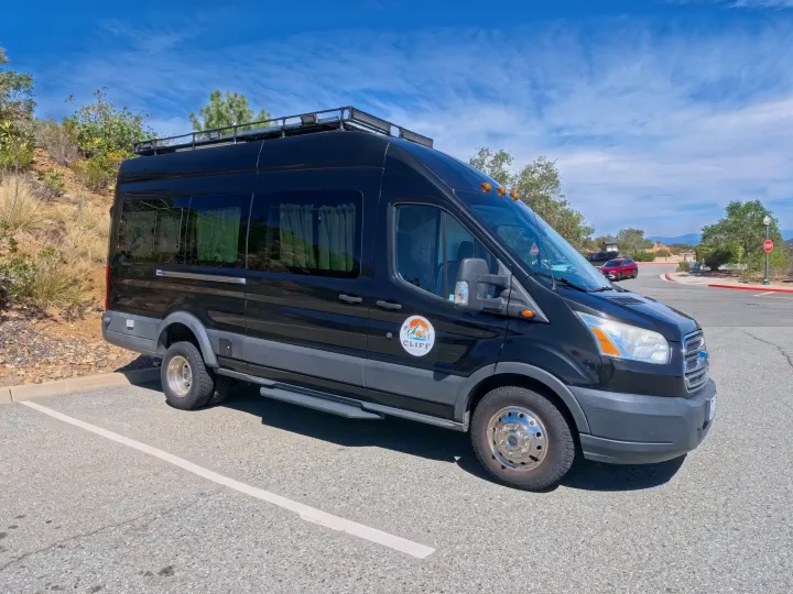 Black Ford Transit campervan RV rental with a roof rack, parked outdoors under a blue sky, ready for travel adventures.. Ford Transit Custom 2015