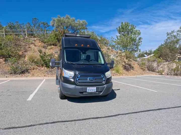 A black Ford Transit camper van RV rental, with a roof light bar, is parked in a sunny lot surrounded by a hillside and trees.. Ford Transit Custom 2015