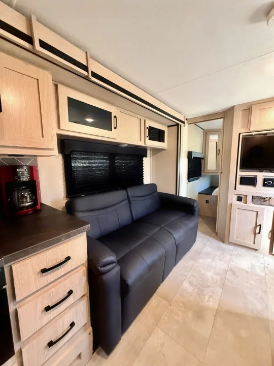 Modern rv rental interior with a black sofa, light wood cabinets, kitchenette with coffee maker, and entertainment area.. Coachmen Apex 2022