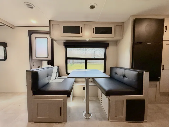 Cozy RV rental dinette with black seating, light cabinets, table, and a refrigerator, ideal for travel.. Coachmen Apex 2022