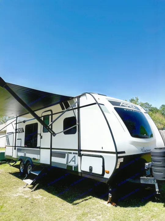 A white and black travel trailer with an extended awning on a sunny, grassy lawn. An ideal RV rental for outdoor adventures.. Coachmen Apex 2022