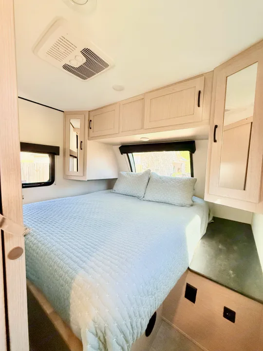 Modern RV rental bedroom with light wood cabinets, a comfy quilted bed, window, and ceiling AC unit.. Coachmen Apex 2022