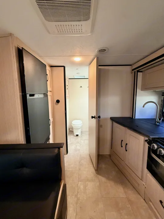 This RV rental interior features a modern kitchen with sink and stove, a large refrigerator, sofa seating, and a compact bathroom with a toilet.. Coachmen Apex 2022