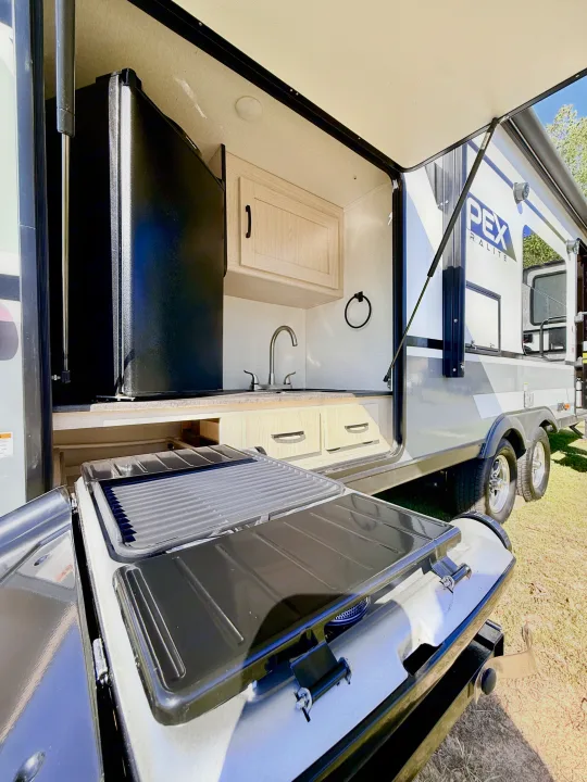 An RV rental's outdoor kitchen with a pull-out grill, refrigerator, and sink. Perfect for convenient camping and outdoor cooking.. Coachmen Apex 2022
