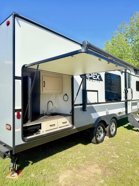 An Apex Ultralite RV rental displays its outdoor kitchen, complete with a sink, cabinets, fridge, and pull-out grill.. Coachmen Apex 2022